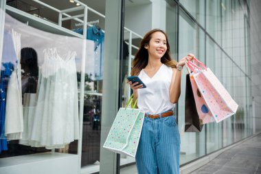 Asian beautiful woman holding shopping bag and smartphone on her hands near the mall shop window, Smiling shopper young girl standing using mobile phone after shopping on city street