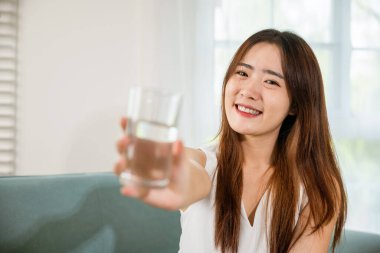 Oturma odasında su bardağıyla gülümseyen mutlu kadın güzelliği, evde içme suyu tutan Asyalı genç kadın portresi, sağlıklı yaşam tarzı.