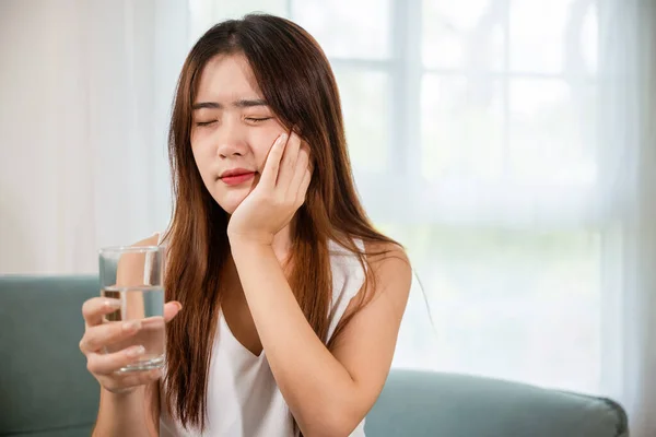 冷たい水を飲んだ後の頬の痛みに触れる美しい女性の歯痛と歯科の問題 自宅のリビングルームで冷たい水のガラスを保持敏感な歯を持つアジアの若い女性 — ストック写真