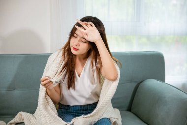 Hasta Asyalı genç kadın kanepede battaniyenin altında oturup evdeki oturma odasında, soğuk algınlığı ve migren konsepti, tıbbi sağlık hizmetleri, dijital termometre ve kağıt ile sıcaklığı ölçerek hapşırıyor.
