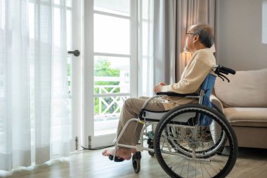 Asian senior man disabled sitting alone in wheelchair looking through window at hospital, lonely elder thoughtful sad old man look outside windows in bedroom at retirement home, feel depressed lonely