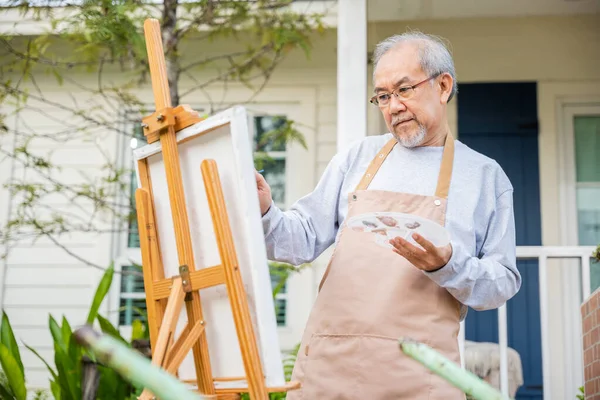 Lifestyle elderly people smile paint at his easel outside home, Asian senior old man painting picture using brush and oil color on canvas, Happy retirement artist and activity concept