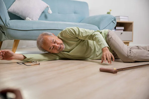 Asian old man lying on floor after falling down with wooden walking stick, Sick senior man beside couch on rug in living room at home, Elderly having an accident after doing physical therapy alone