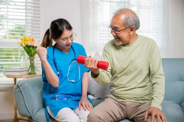 若い看護師理学療法士は 自宅でダンベルを持ち上げるのに役立ちます アジアの看護師は 赤いダンベルワークアウトを使用してソファの上に座っている高齢者の退職をサポートします運動物理的な回復 ヘルスケアの概念 — ストック写真