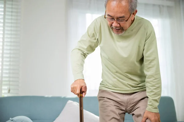 眼鏡をかけたアジアの老人は 杖でソファから立ち上がる 膝の痛みに苦しむ高齢者杖のハンドルを握る 老人は家で杖を握る — ストック写真