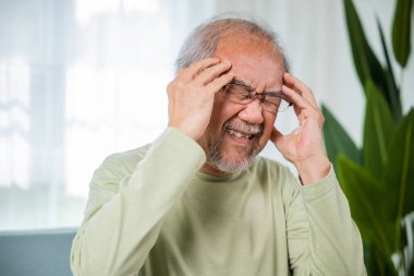 Baş ağrısı. Migren ağrısından muzdarip yaşlılarla el ele tutuşan üzgün Asyalı son sınıf öğrencisi koltukta oturmuş evde yalnız ve yaralı hissediyor yaşlılık sağlık sorunları, sağlık sorunları...
