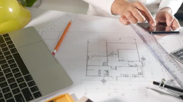 建築プロジェクトの職場 スマートフォンや携帯電話を使って家のデザインをレビューする建築家は 設計図の鉛筆で図面を編集する前に 建築事務所の机の上の設計図の紙 — ストック動画