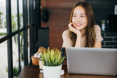 Dışarıda çalışan, kahvecide gülümseyen, serbest çalışan Asyalı genç iş kadını, kafeteryada tek başına oturmuş, camdan dışarı baktığında bilgisayarını gören mutlu bir kadın.