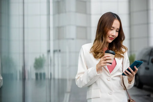 コーヒーカップを持っているアジアの実業家は スマートフォンが午前中にオフィスの近くの街の通りを歩く仕事に行く見て 肖像画のビジネス女性は携帯電話とホットドリンク屋外の紙カップを保持します — ストック写真