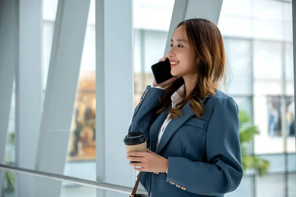 Asian business young woman confident hold drinking coffee takeaway to go and talking on mobile phone, happy female with coffee cup calling on smartphone in street near modern office