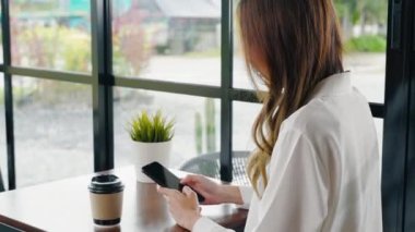 Pencerenin yanındaki kafede oturan mutlu bir kadın cep telefonu, Asyalı genç iş kadını serbest telefon ve kahve dükkanında kahve kağıdı bardağı tutuyor.