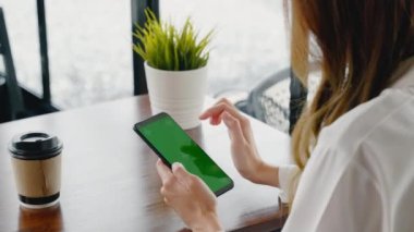 Pencerenin yanındaki kafede oturan mutlu bir kadın cep telefonu yeşil ekranda, Asyalı genç iş kadını serbest telefon ve kahve fincanında kahve kağıdı tutuyor.
