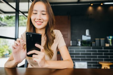Asyalı genç bir kadın, sabah kafede online alışveriş yapmak için akıllı telefon kullanıyor. Kadın, cep telefonundan para transferi yapıyor.