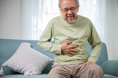 Elderly Asian old man sitting on sofa having stomachache, old age suffering from stomach ache holding his stomach in living room, people health problem, food poisoning clipart