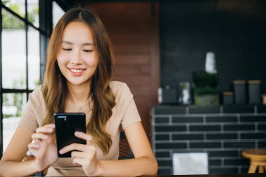 Güzel Asyalı kadın el ele tutuşup diğerleriyle cep telefonuyla iletişim kurmak için daktilo kullanıyor. Genç kadın sabahları pencerelerin yanındaki kafede online alışveriş yapmak için akıllı telefon kullanıyor.