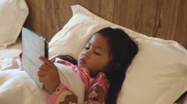 Little Asian girl playing digital tablet on bed addicted game and cartoon, Kid watching videos and cartoons online in bedroom at home, Fun online concept