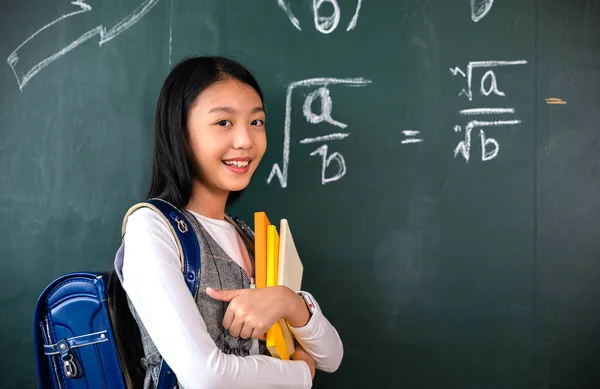 Back to school. Happy beautiful Asian Schoolgirl girl standing holding books standing in front blackboard of classroom, Portrait of smiling woman child student of black chalk board, Education lesson
