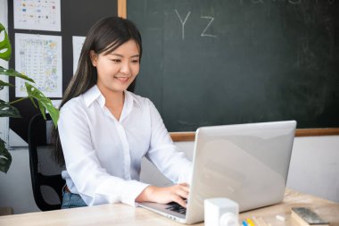 Okula dönelim. Okul masasında bilgisayar kullanan gülümseyen kadın, sınıfta dizüstü bilgisayarı olan genç kadın öğretmen portresi, online eğitim ve öğrenim konsepti.