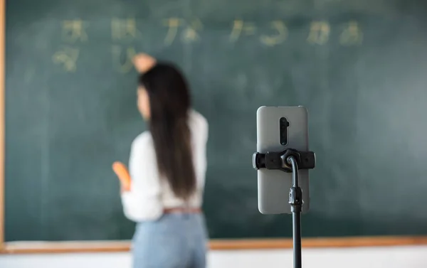 Back to school online class. Asian teacher teach on board while have blackboard lecture online through smartphone in classroom, woman standing front of class live video call online smart mobile phone