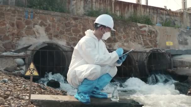 生态学家从检查清单中抽取有毒化学品的水 并写信给剪贴板纸 生物学家穿着防护服 戴着面具 从工业废水中采集样本 — 图库视频影像