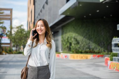 Asyalı iş kadını akıllı cep telefonundan arıyor ve ofis binasının önünde caddede yürüyor, şehirde akıllı telefondan konuşan mutlu iş kadını, müşteriyle iş iletişimi...