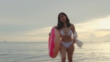 Genç, güzel bir kadın elinde lastik yüzükle tropikal sahilden yürürken bikini giyer, tatilde yüzdükten sonra şişme yüzük takar yaz tatili yapar.