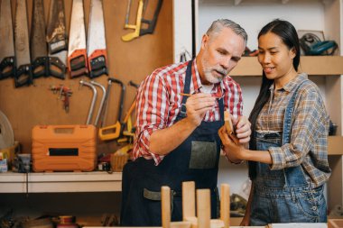 Esnaf kıdemli adam marangozhanede kadın çırak eğitiyor, marangoz atölyede ahşap masa başında duran kadın için ahşap eğitiyor, Ulusal Marangozlar Günü.