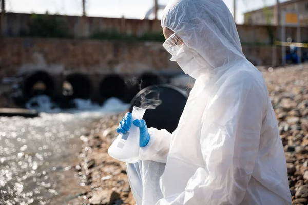 Ecologist sampling water toxic chemicals from river with test tube glass and have white smoke, Biologist wear protective suit and mask collects sample waste water from industrial, problem environment
