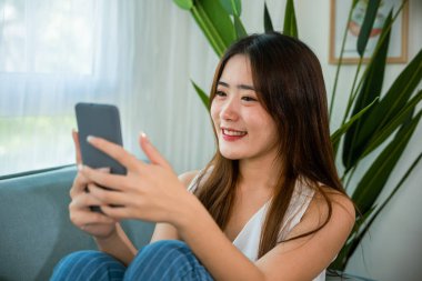 Sosyal medyayı kontrol eden mutlu Asyalı genç bir kadın elinde akıllı telefonuyla kanepede dinlenirken, güzel bir kadın kanepede oturmuş internetten alışveriş yaparken gülümsüyor.