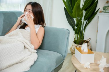 Asian young woman she sick blowing nose sneezing in tissue at home, fever caught cold, Sick female sitting under blanket on sofa and sneeze with tissue paper in living room, medicine healthcare