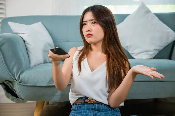 Unhappy young woman watch movie on television at home, bored beautiful female holding tv remote control sitting floor of sofa in living room at home, boring with television program