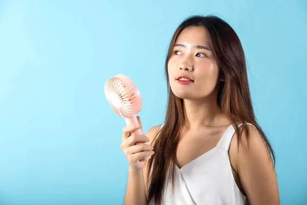 Asian beauty young woman holding pink portable electric mini fan near her face studio shot isolated on blue background, Female hand hold small plastic fan handheld she enjoying cool wind blowing