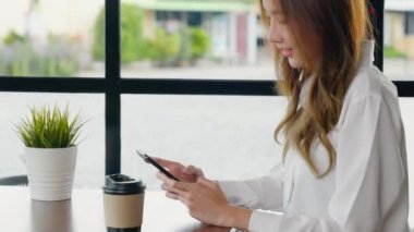 Pencerenin yanındaki kafede oturan mutlu bir kadın cep telefonu, Asyalı genç iş kadını serbest telefon ve kahve dükkanında kahve kağıdı bardağı tutuyor.