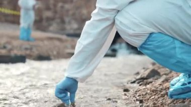 Nehirdeki suyu test tüpüyle örnekleyen ekolojist, koruyucu kıyafet giyiyor ve maske endüstriyel, sorunlu çevreden ve bilim adamı çevreciden atık su örnekleri topluyor.