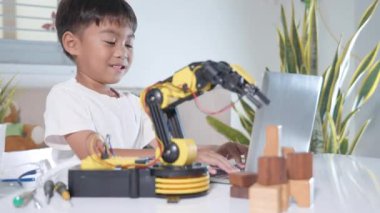 Asyalı küçük çocuk atölyede dizüstü bilgisayar kullanıyor. Robotik kol makinesini kontrol ediyor. Programlama kontrol robotu kolu için çocuk öğrenme programcısı.