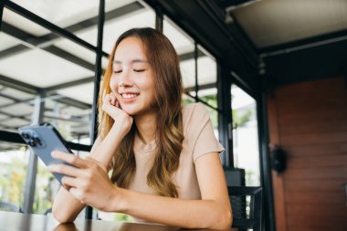 Güzel Asyalı kadın el ele tutuşup diğerleriyle cep telefonuyla iletişim kurmak için daktilo kullanıyor. Genç kadın sabahları pencerelerin yanındaki kafede online alışveriş yapmak için akıllı telefon kullanıyor.