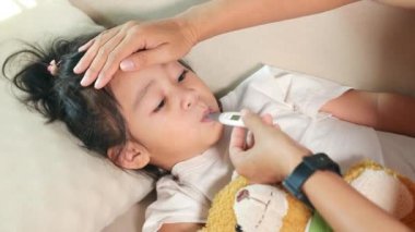 Sick kid. Mother parent checking temperature of her sick daughter with digital thermometer in armpit, child laying in bed taking measuring her temperature for fever and illness, healthcare