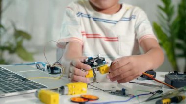 Asyalı çocuk tekeri Arduino robot araba ev ödevine monte ediyor, küçük çocuk tekerleği araba oyuncağına monte etmeye çalışıyor, elektronik yapay zeka atölyesi online okul dersi yaratıyor.