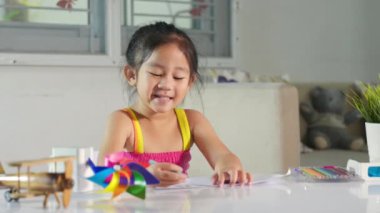 Masaya oturmuş gülümseyen Asyalı sevimli çocuk evde resim çiziyor ve resim çiziyor sanat ödevi öğreniyor, küçük mutlu kız çocuğu boyamadan önce kağıda karikatür çiziyor.