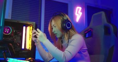 You win. Excited Asian woman gamer wearing gaming headphones playing joystick console video game and streaming online on computer PC neon light at home happy successful, tournament player E-Sport