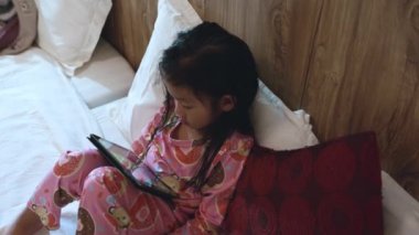 Küçük Asyalı kız yatak bağımlısı oyun ve çizgi film üzerinde dijital tablet oynuyor, çocuk yatak odasında video ve çizgi film izliyor, online eğlence kavramı var.