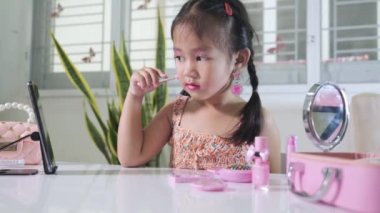 Makyaj yapan Asyalı sevimli, komik küçük kız pudra için akıllı telefon ve yanak fırçasına bakıyor, kadın olmak için etkinlik öğreniyor, sosyal medya blogcusu kozmetik ile vlog kaydediyor.
