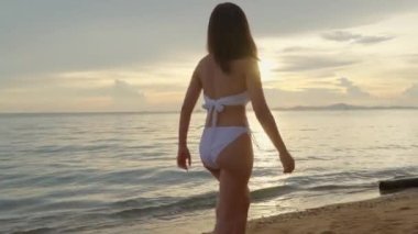 Arkaya bakış, genç güzel bir kadın bikini giyiyor tropikal sahilde yürüyor ve altın gün batımında özgürlük için elini kaldırıyor mutlu Asyalı kadın gün batımında tatilde dinleniyor yaz tatili