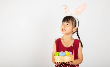 Mutlu Paskalyalar. Paskalya tavşanı kulakları takan gülümseyen Asyalı küçük kız elinde renkli yumurtalarla dolu bir sepet beyaz arka planda izole edilmiş bir şekilde gülümsüyor.