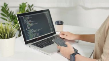 Programmer woman looking mobile phone program code at workplace, software developer hands typing data coding programming javascript with laptop computer, IT startup concept, Web application design