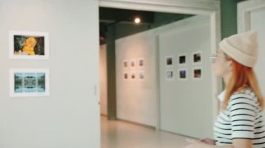 Asian woman walking through art gallery collection in front framed paintings pictures on white wall, Visitor person walk looking to picture at photo frame at interior show exhibition artwork gallery