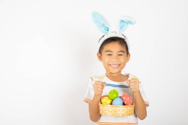 Mutlu Paskalyalar. Paskalya tavşanı kulakları takan gülümseyen Asyalı küçük çocuk elinde renkli yumurtalarla dolu bir sepet beyaz arka planda izole edilmiş bir şekilde gülümsüyor.