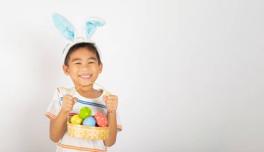 Mutlu Paskalyalar. Paskalya tavşanı kulakları takan gülümseyen Asyalı küçük çocuk elinde renkli yumurtalarla dolu bir sepet beyaz arka planda izole edilmiş bir şekilde gülümsüyor.