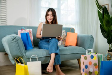 Asyalı genç bir kadın alışveriş poşetleriyle kanepede oturuyor bilgisayarla online alışveriş yapıyor, mutlu bayan müşteri evdeki oturma odasındaki web sitesinden satın almakla tatmin oluyor.