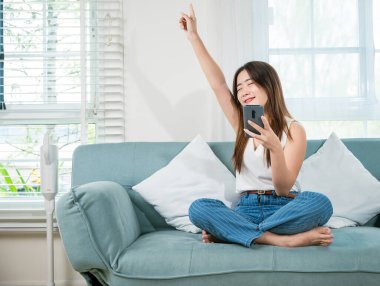 Yaşam tarzı, kablosuz kulaklıklı bir kadın müzik dinlemeli bir şarkı ve müzik uygulamalı akıllı telefondan biraz dans, Asyalı bir kadın oturma odasında akıllı cep telefonuyla kanepede müzik dinlerken kulaklık takıyor.